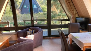 Ein gemütliches Zimmer mit einer Couch, einem Tisch und einem Fernseher. Große Fenster bieten Blick auf ein Holzhaus und umliegende Grünflächen.