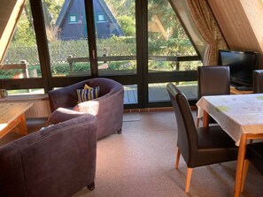 A cozy room with a sofa, a table, and a TV. Large windows offer a view of a wooden house and greenery outside.