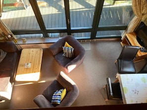 Une pièce avec une table basse en bois, deux fauteuils avec des coussins, une petite table avec un clavier, et une vue sur une terrasse extérieure, visible à travers de grandes fenêtres.