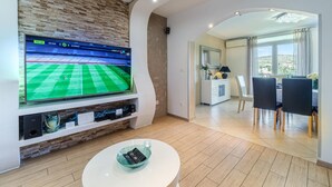 A modern living room with a large TV displaying a sports event, a white coffee table, and a dining area with a view of the outdoors. - Amazing home in Milna (Milna)