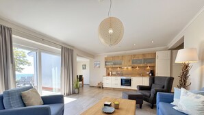 Een moderne woonkamer met een bank, fauteuil en salontafel. Er is een keuken met houten kastjes en een inbouwoven. Een groot raam biedt uitzicht op het buitengebied.