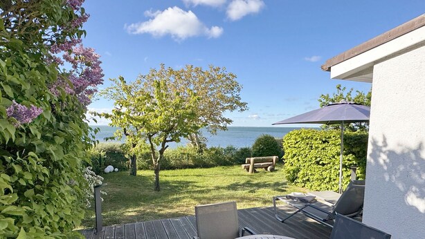 Een terras met tafel en stoelen, een houten terras, uitzicht op zee en een tuin met bomen en bloemen.