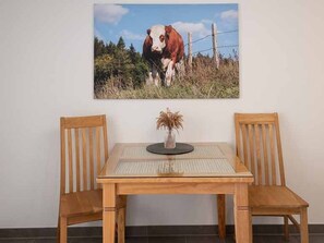 Una mesa de comedor con dos sillas de madera, un florero con plantas secas y un cuadro con una vaca en la pared.