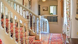 Un magnifique escalier orné de balustres travaillés, une moquette rouge à motifs et une vue sur une pièce dotée d’une grande fenêtre et d’un tableau accroché au mur.