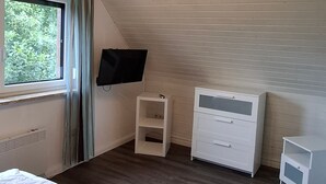 A bedroom with a sloped ceiling, a bed, a TV, a small white cabinet, and a nightstand. - Vacation cottage Sander, Germany (Friedrichskoog)