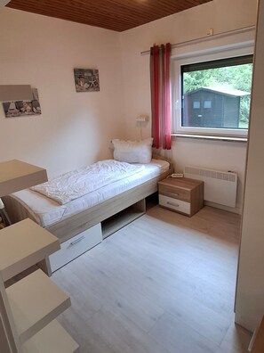A small, single-bed room with a wooden ceiling, a window with red curtains, a radiator, and a small bedside table. - Vacation cottage Sander, Germany (Friedrichskoog)