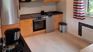 A compact kitchen with wooden cabinets, a stainless steel refrigerator, a microwave, an oven, a dishwasher, and a coffee maker on the counter. - Vacation cottage Sander, Germany (Friedrichskoog)
