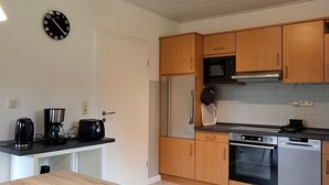 A modern kitchen with wooden cabinets, a stainless steel refrigerator, an oven, and a microwave. There is a clock on the wall and a coffee maker on the counter. - Vacation cottage Sander, Germany (Friedrichskoog)