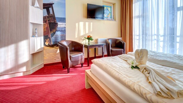 Quarto de hotel com cama, duas cadeiras, uma mesinha, televisão e um mural de um navio na parede.