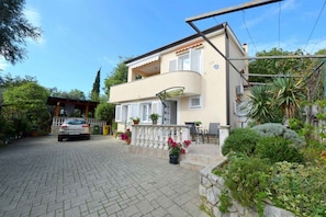 A two-story house with a balcony, a car parked in front, and a garden with various plants.