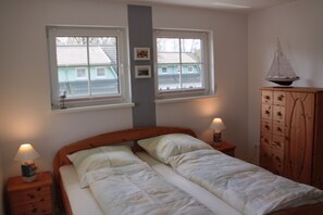A bedroom with a double bed, two bedside tables with lamps, a wooden wardrobe, and two windows with a view of houses.