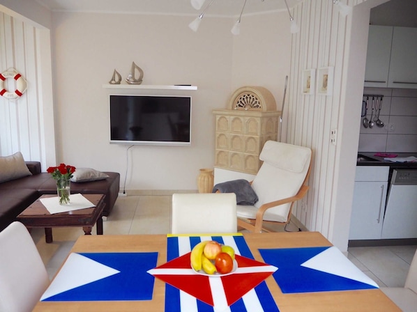 A modern living room with a flat-screen TV, a sofa, a dining table with a flag design, and a kitchen area with cabinets and utensils.