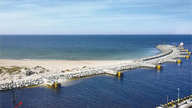 Ein Küstensteg, der sich ins Meer erstreckt, mit einem Sandstrand und Felsformationen in der Nähe.