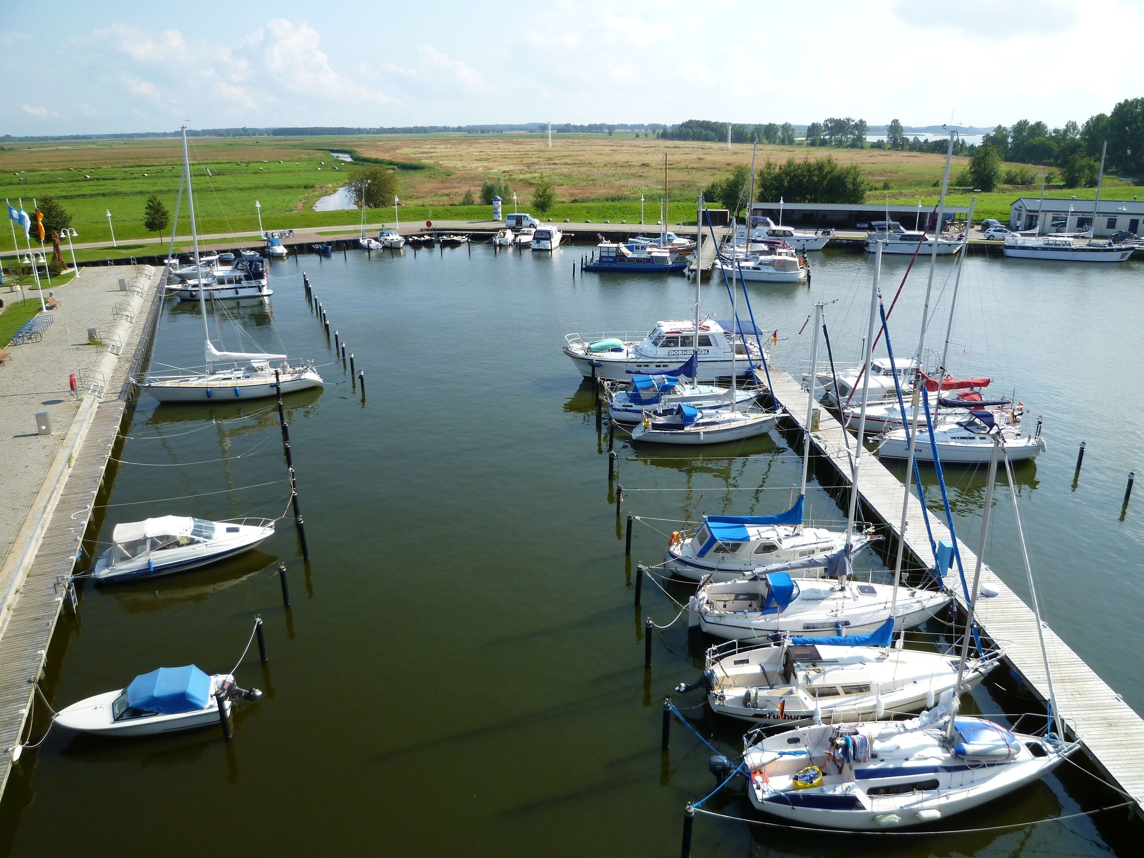 A marina with various boats docked, a grassy field in the background, and a clear sky.