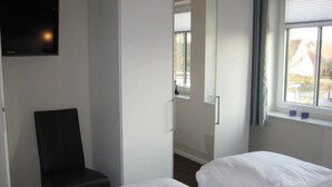 A hotel room with a bed, a chair, a TV, and a window with blinds. - 2-room 103 Juist Balk Spülm. (BC 5) - BUE - Apartment house Westwind (Büsum)