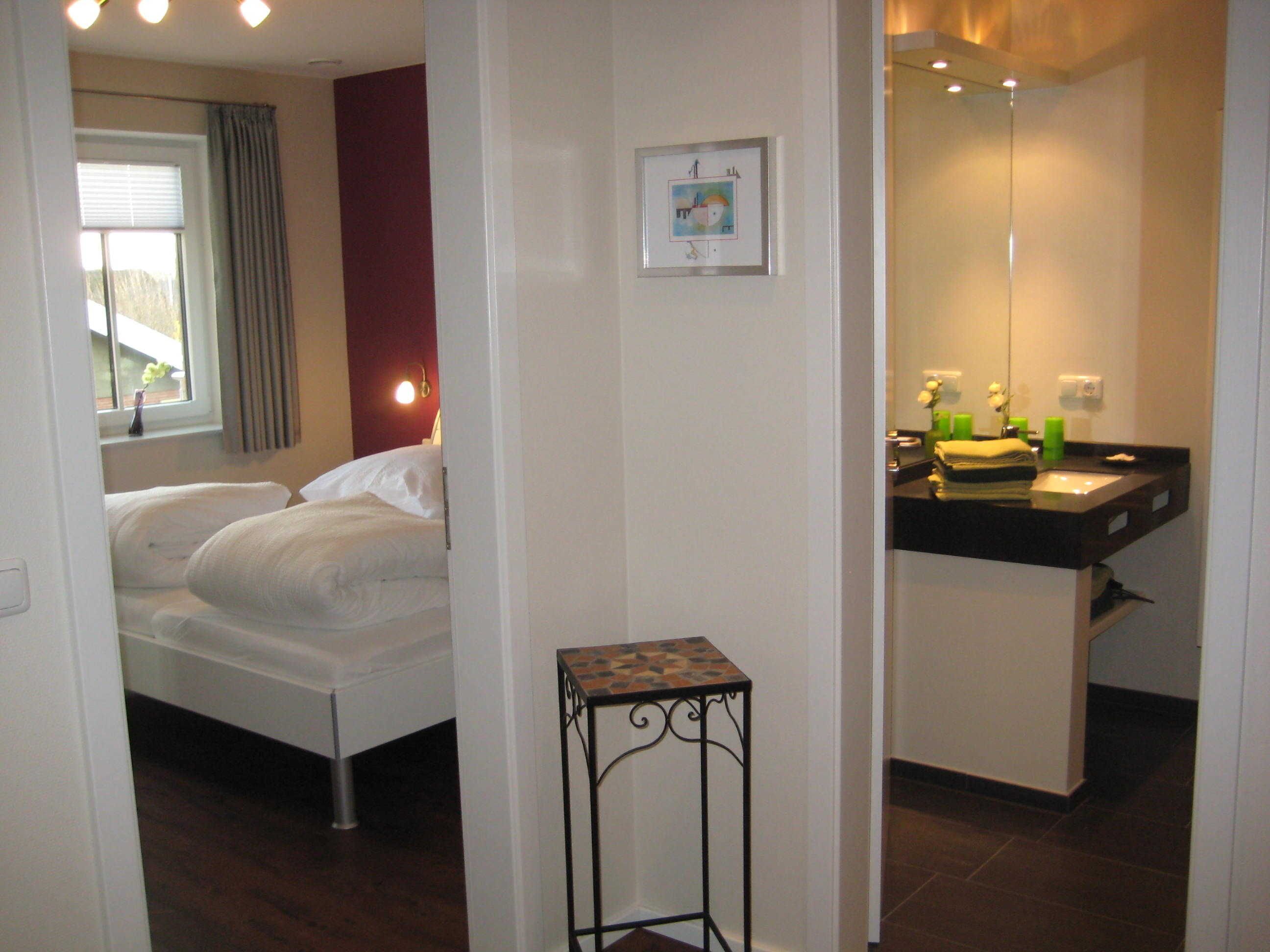Un dormitorio con cama, mesita de noche, ventana con cortinas y baño con lavamanos y espejo.