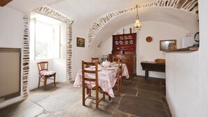 Un comedor con suelo de piedra, sillas de madera, una mesa con mantel, un armario rojo con vajilla y una lámpara colgante.