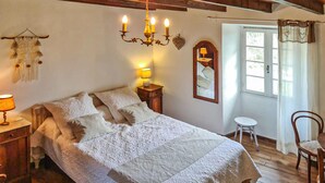 Un dormitorio con una cama grande, una mesita de noche con lámpara, un espejo, un taburete pequeño y una ventana con cortinas.
