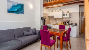 Um espaço compacto com cozinha compacta, área de jantar com mesa de madeira e cadeiras roxas, sofá cinza e uma obra de arte na parede retratando uma onda azul.