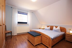 Ein Schlafzimmer mit einem hölzernen Bett, einem Nachttisch, einem Sessel, einem Fenster mit Vorhängen und einer Heizung.