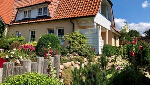 Une maison à deux étages avec un toit recouvert de tuiles rouges, une porte de garage blanche et un jardin où poussent différentes plantes et fleurs.