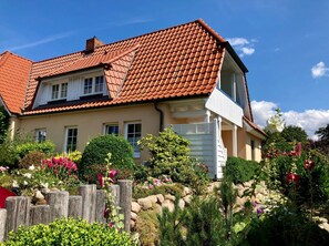 Une maison à deux étages avec un toit recouvert de tuiles rouges, une porte de garage blanche et un jardin où poussent différentes plantes et fleurs.