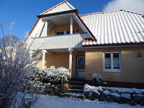 Een tweelaags huis met een balkon, een sneeuwbedekt dak en een stenen muur ervoor.