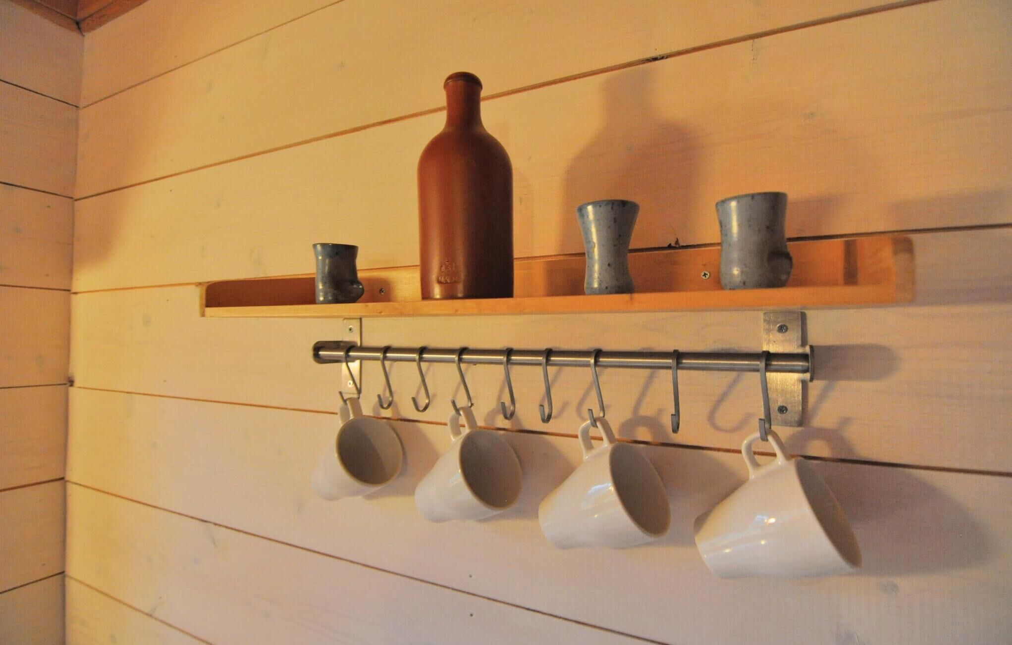 A kitchen shelf with a brown bottle, two metal cups, and a metal hook rack holding white cups.