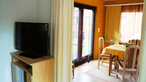 A room with a television on a wooden stand, a dining table with chairs, and a sliding glass door leading to an outdoor area.