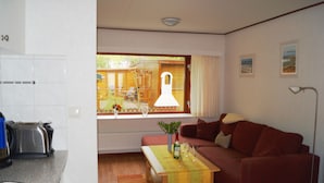 A compact living space with a kitchenette, a red sofa, a dining table with a bottle and glasses, and a window with a view of a wooden deck and outdoor furniture.