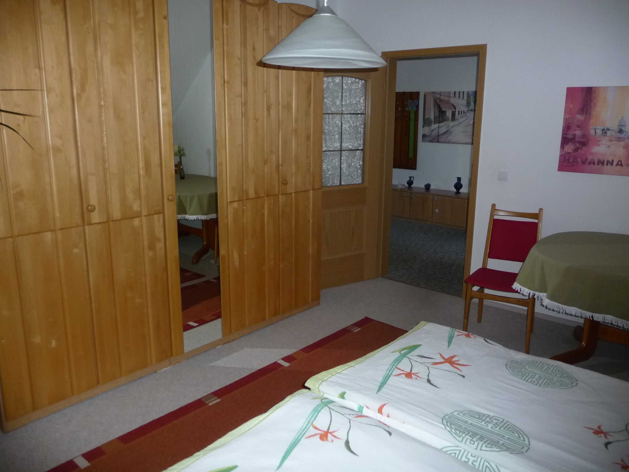 A room with a bed, a chair, a table, a hanging lamp, and a door leading to another room.