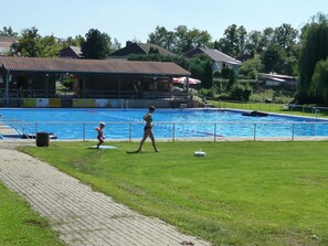 Une piscine avec une pelouse devant, un chemin pavé et des maisons en arrière-plan.