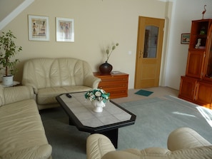 A living room with a sofa, a coffee table with a vase, a wooden cabinet, and a door. - Apartment OG - Apartment / DZ Blesse Allstedt (Allstedt)