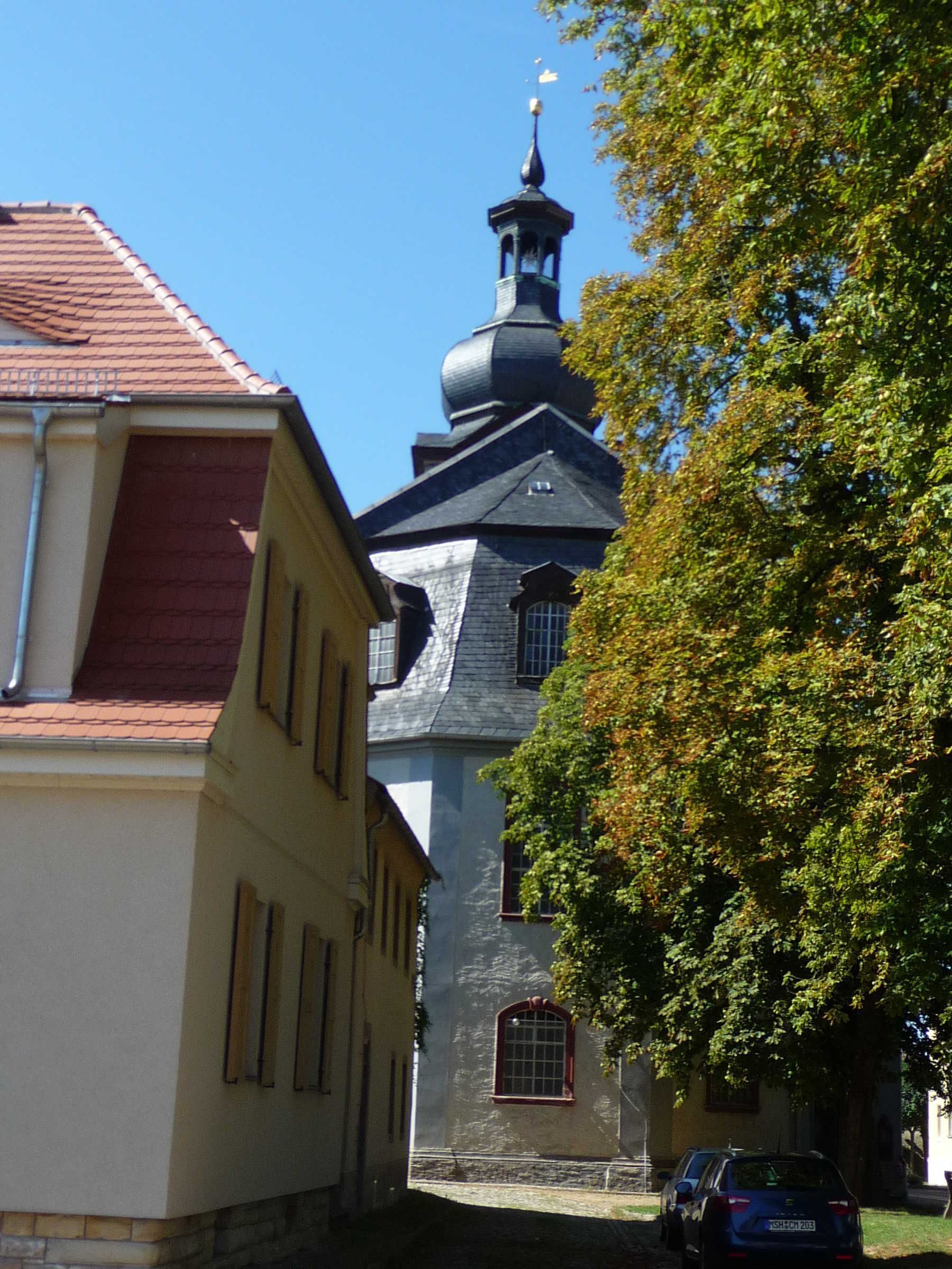 A narrow street with a church tower, a parked car, and a tree.