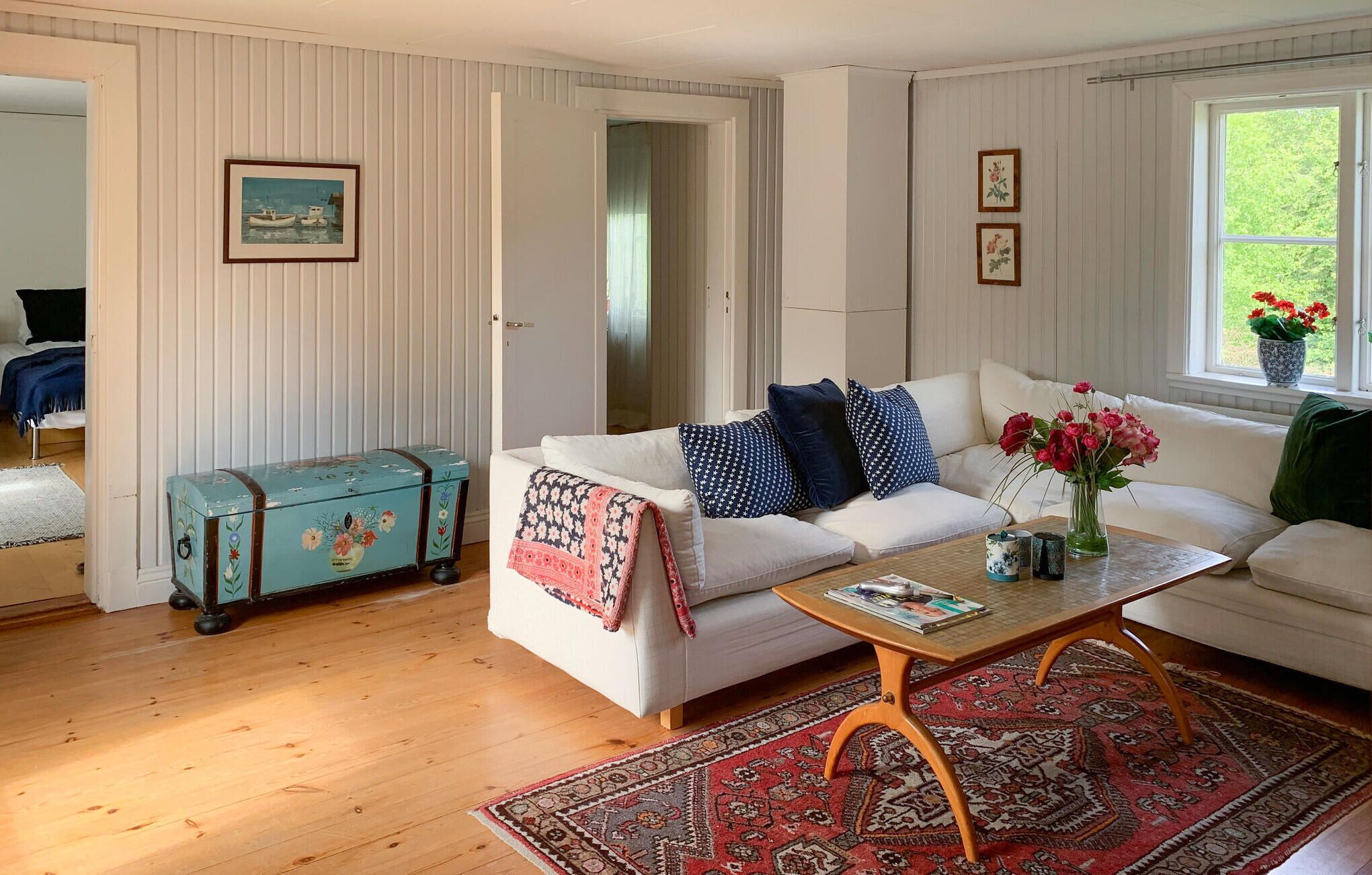 A living room with a white sofa, a patterned throw blanket, a vintage suitcase, a coffee table, and a large window with a view of greenery.
