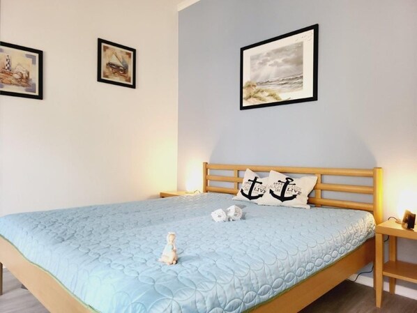 A neatly made bed with a blue quilt, two framed artworks on the wall, and a wooden nightstand with a lamp.