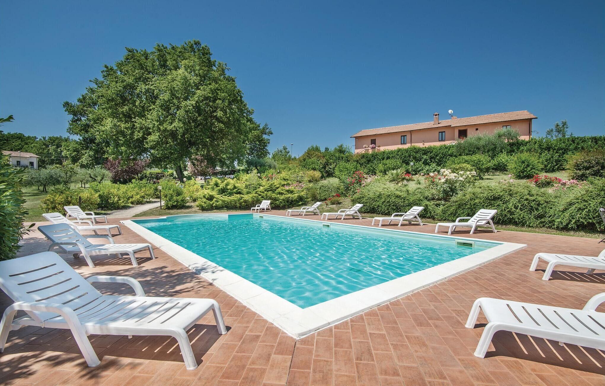 A rectangular swimming pool with white poolside chairs, surrounded by a brick patio, with a large tree and a building in the background.