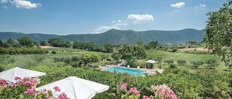 A pool with lounge chairs and umbrellas, surrounded by greenery and mountains in the background.