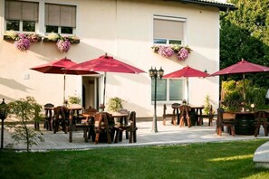 Un patio al aire libre con mesas y sillas de madera, sombrillas rojas y cestas de flores colgantes.