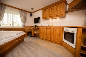 Une chambre avec un lit en bois, un bureau en bois, une chaise, une télévision et un coin cuisine doté de meubles de rangement en bois et d’un four.