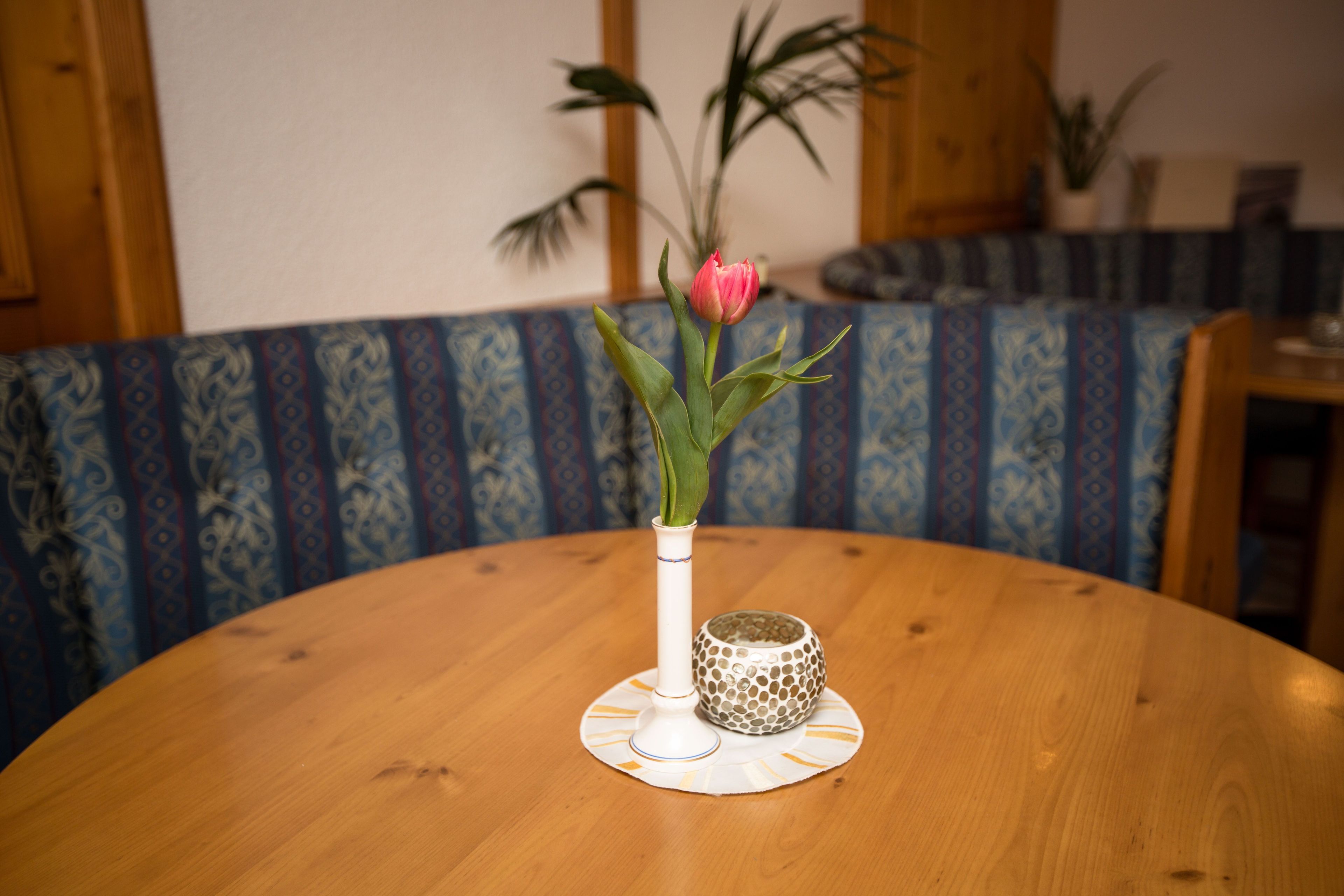 Ein runder Holztisch mit einer weißen Vase, einer rosa Blume, einem gemusterten Kissen und einer dekorativen Schale.
