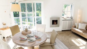 A dining area with a round table set for four, surrounded by chairs. There is a large window with a view of greenery outside. - Residence Störtebeker Whg. FB20-18 .. - Residenz Störtebeker Whg. 18 (Kühlungsborn)