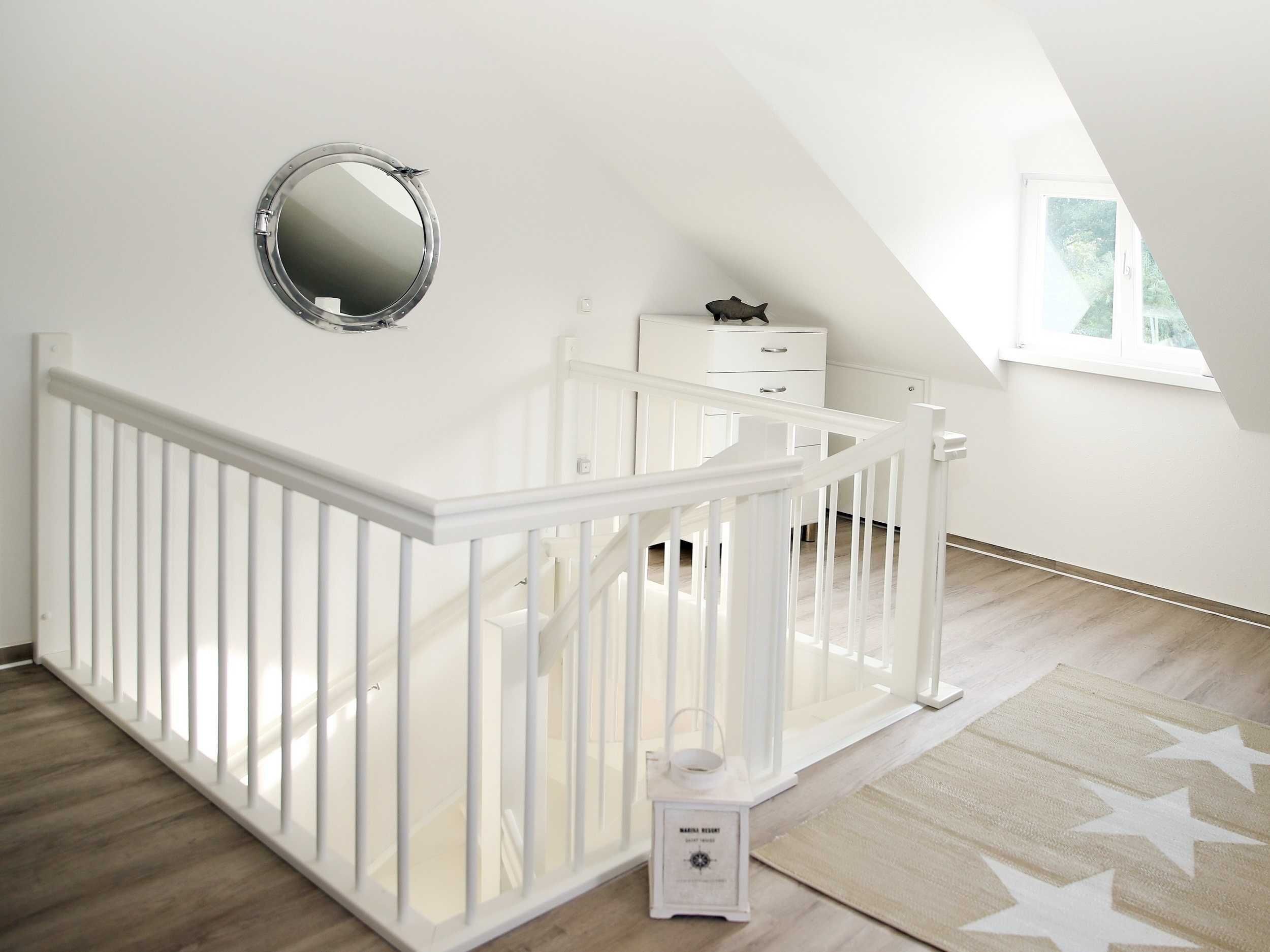 A white crib with a safety rail, a small bedside table, a nightstand, a mirror, and a star-patterned rug in a bright room with a sloped ceiling and a window.