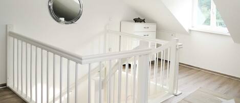 A white crib with a safety rail, a small bedside table, a nightstand, a mirror, and a star-patterned rug in a bright room with a sloped ceiling and a window.