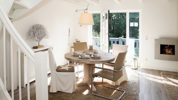 A dining area with a round table, chairs, and a lamp. There is a staircase on the left and a view of the outdoors through large windows. - Residence Störtebeker Whg. FB20-18 .. - Residenz Störtebeker Whg. 18 (Kühlungsborn)