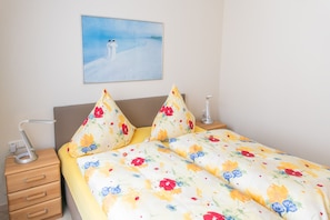 A bedroom with a bed, floral pillows, a bedside table with a lamp, a drawer, and a painting on the wall. - FH Herve, Monica GM 69206-FH Cather (Graal-Müritz)