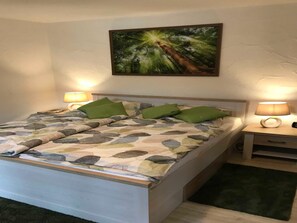 Un lit recouvert d’une couvre-lit à motifs de feuilles, deux oreillers verts, une table de chevet avec une lampe et un tableau représentant une forêt accroché au mur.