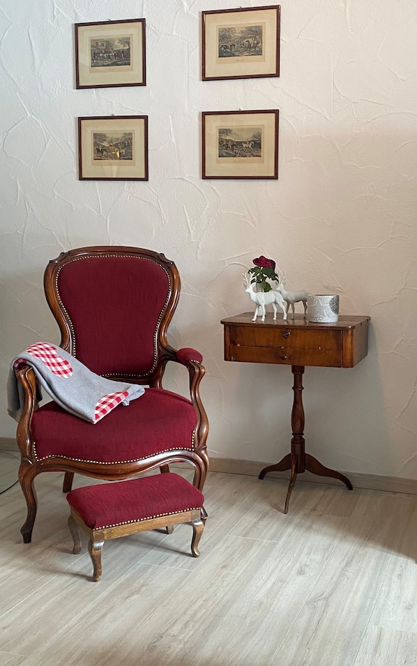 Ein Zimmer mit einem Holzstuhl mit roter Polsterung, einem kleinen Holzhocker mit einer Keramikfigurine und gerahmten Bildern an der Wand.