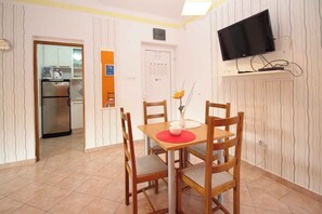 Un comedor con una mesa y sillas, un televisor montado en la pared y una cocina visible a través de una puerta abierta.