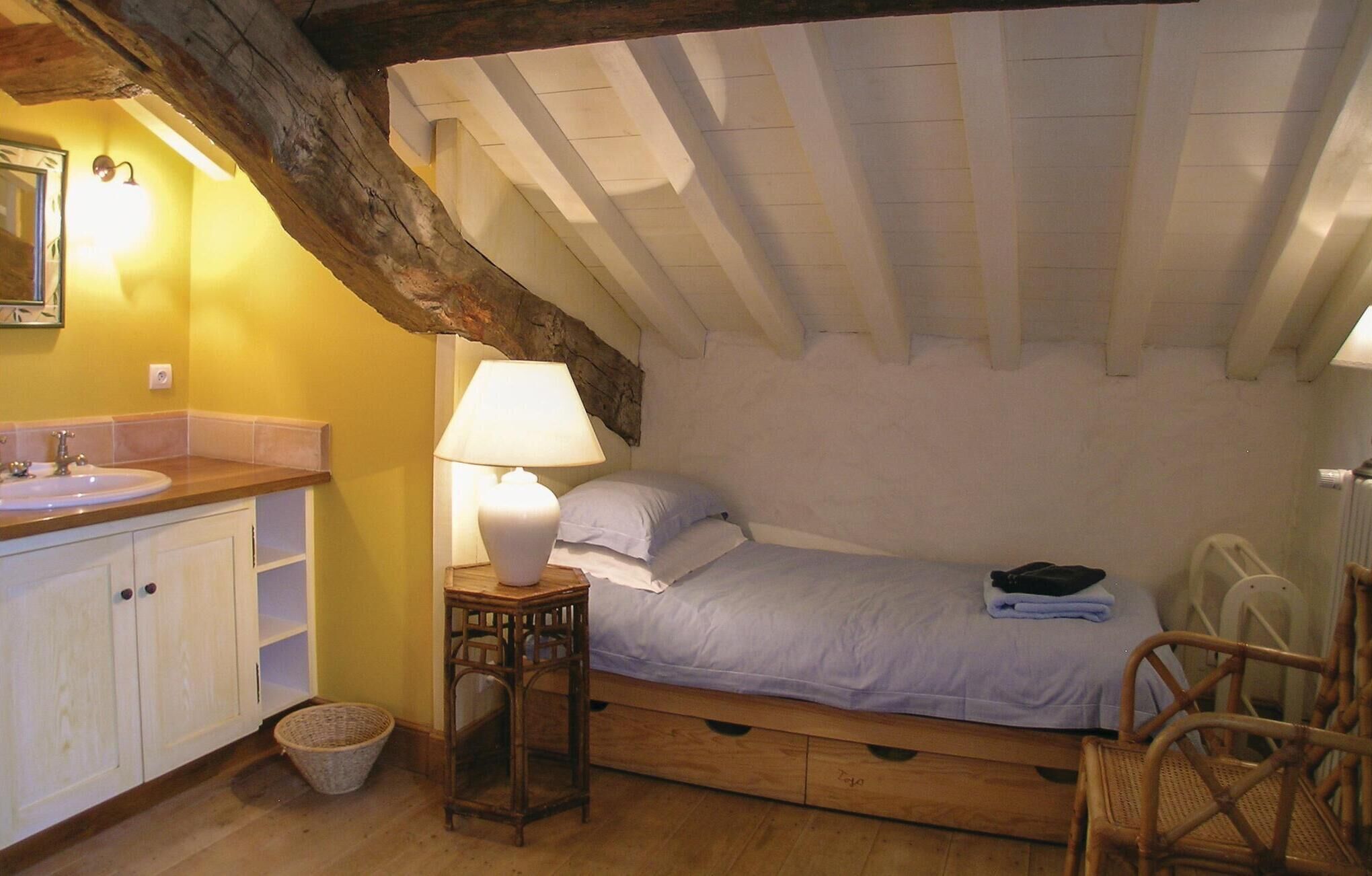Un dormitorio pequeño y acogedor con una cama individual, una mesita de noche con lámpara, una silla de madera y un armario blanco con lavabo.